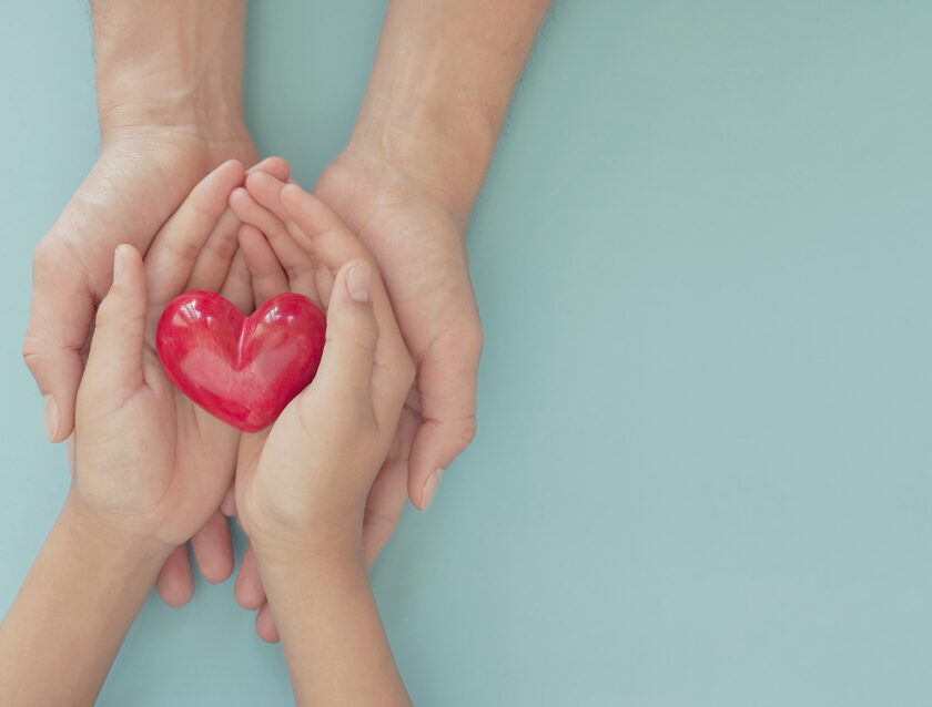 A pair of adult hands and child hands holding a red heart.